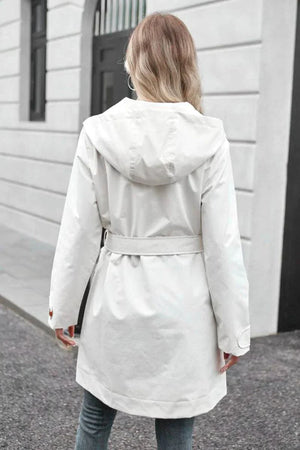 Woman wearing a white water-resistant windbreaker trench jacket with hood and belt, back view