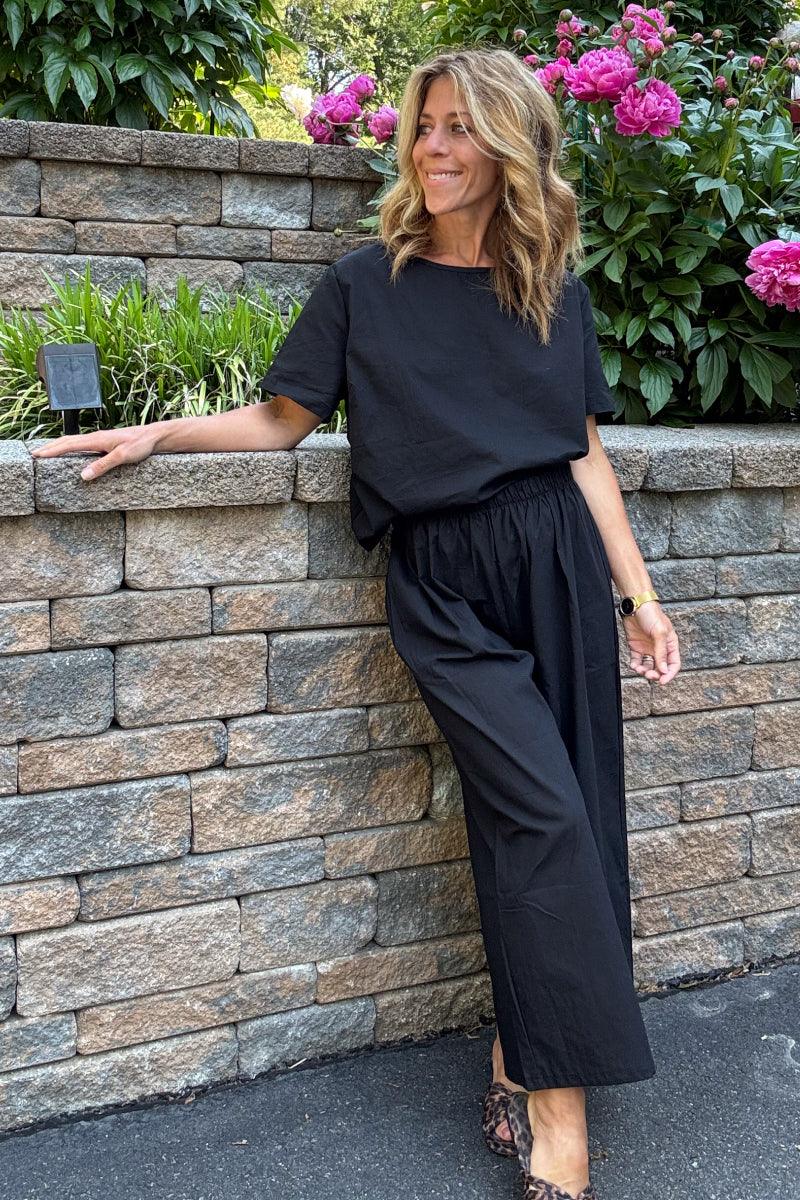 Woman wearing black loose fit co-ord set with short sleeves and wide-leg pants outdoors by stone wall and flowers