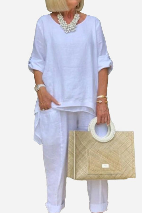 White asymmetrical two-piece trouser set with rolled sleeves and loose fit, paired with a beige handbag and statement necklace
