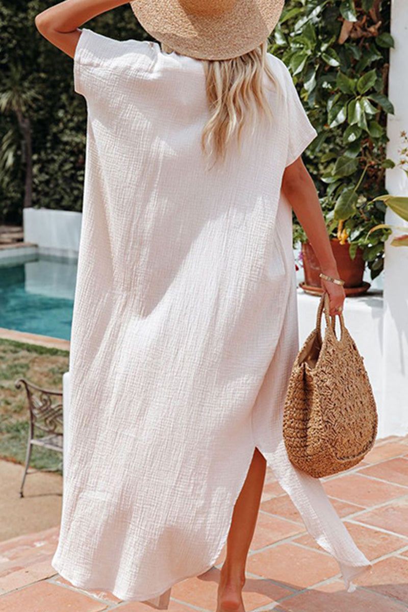 woman wearing a white full-length button-down beach cover-up with a side slit, holding a woven bag and wearing a sun hat by the poolside
