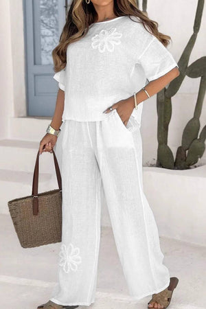 White flowy two piece set with floral embroidery, short sleeves, and wide leg pants, styled with a woven tote bag and casual sandals.