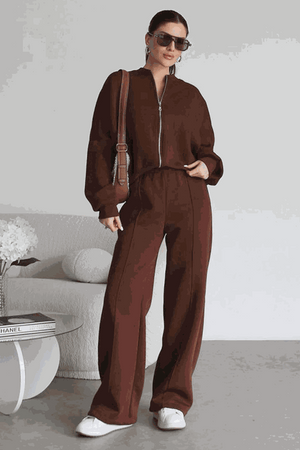 Woman wearing The Voyager Zip Set brown zip-front sweatshirt and wide-leg joggers with pockets, styled with sunglasses and white sneakers in a modern indoor setting
