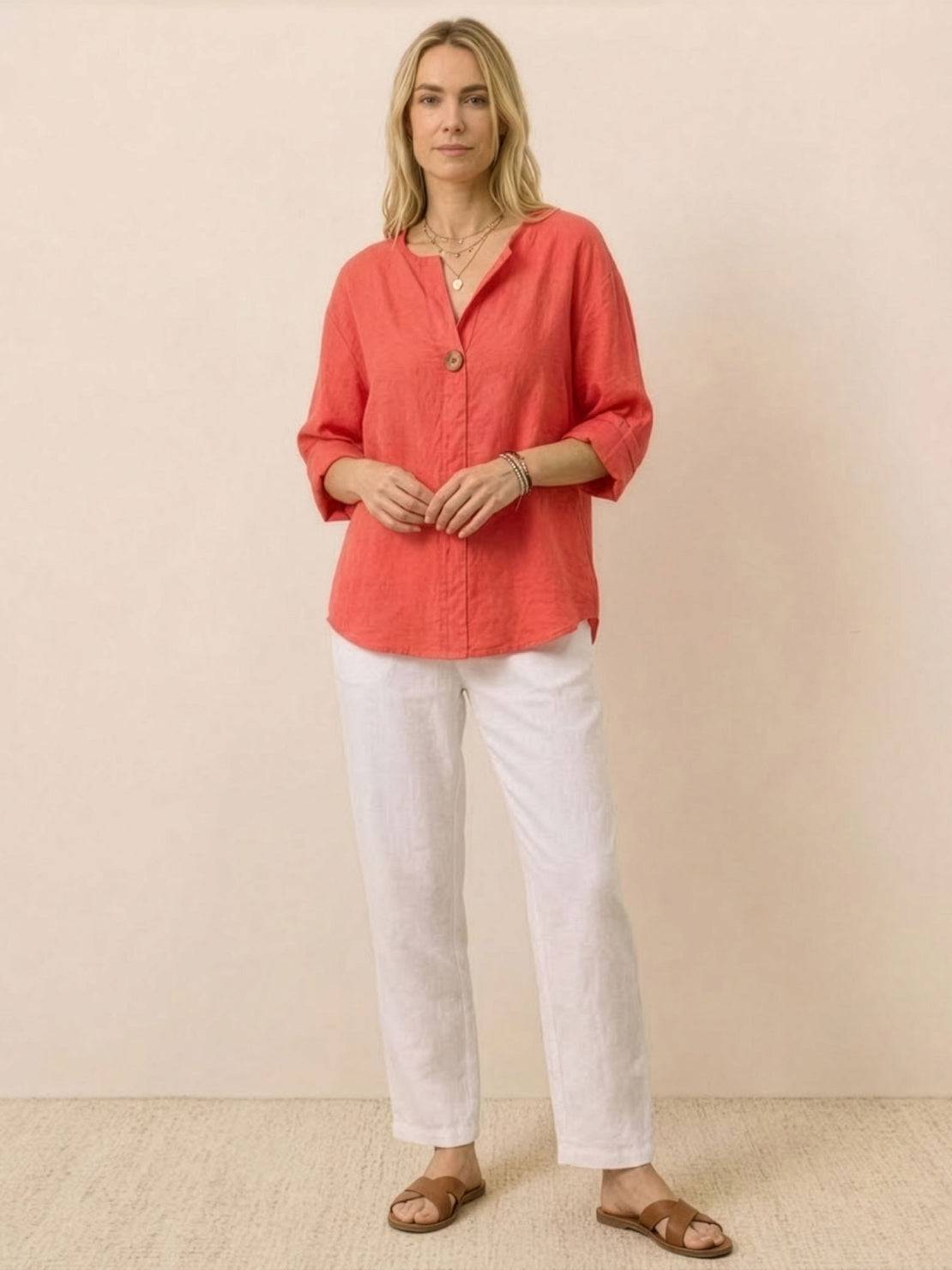 Woman wearing coral pure linen age-defying button detail shirt with white pants and sandals against neutral background