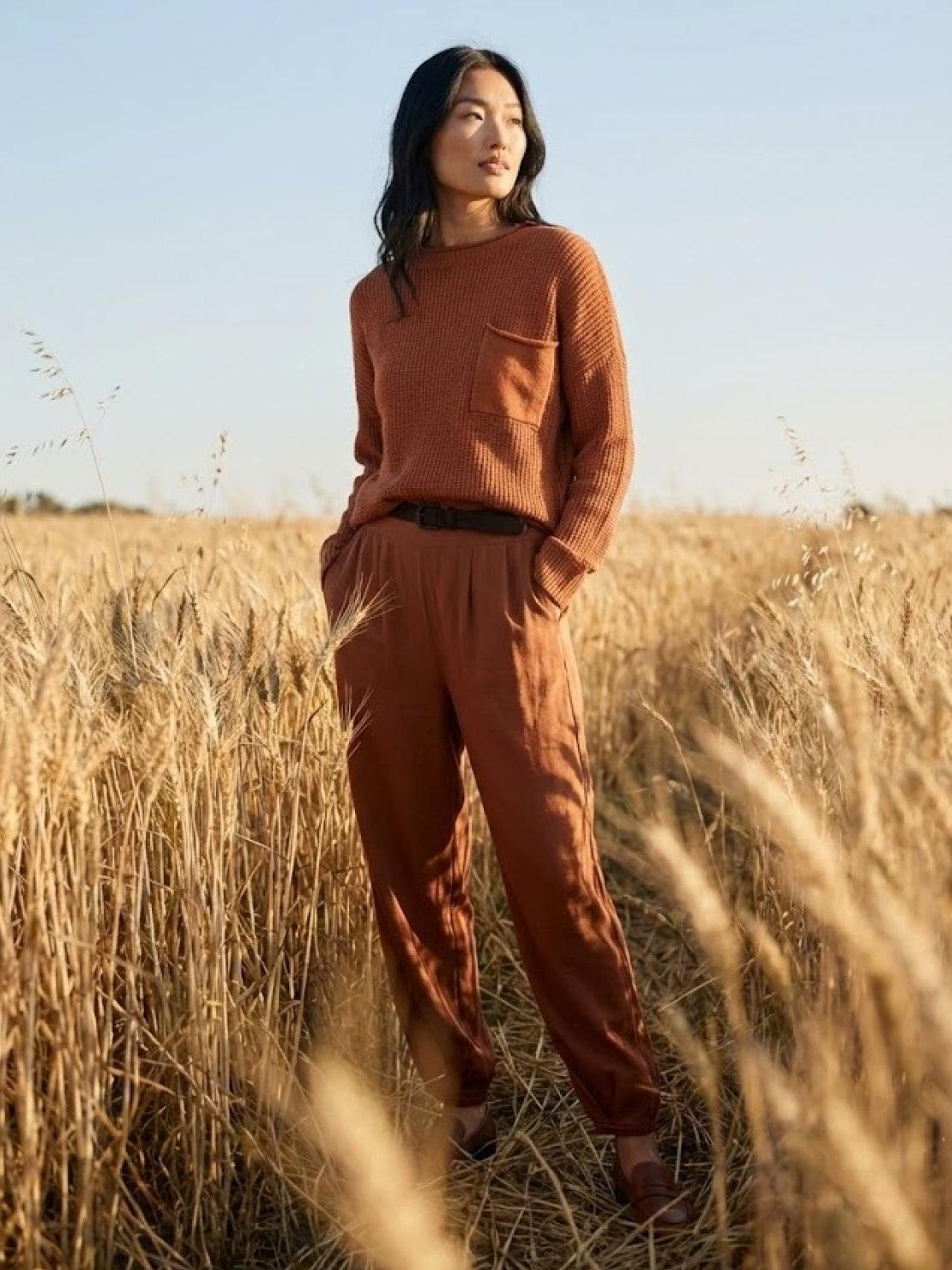 Woman wearing rust-colored relaxed knit sweater top and trousers standing in a wheat field showcasing European minimalism style