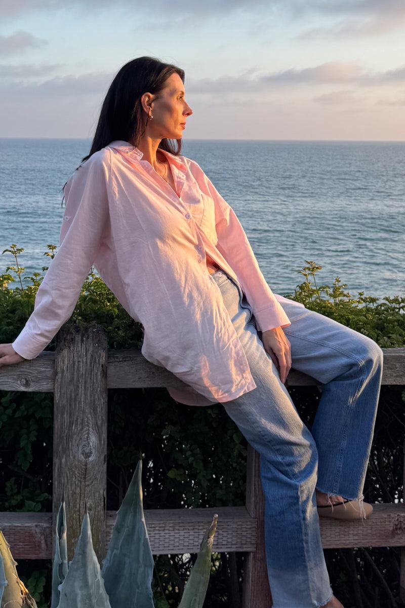 Woman wearing oversized linen shirt in sun-washed pink sitting by the ocean at sunset