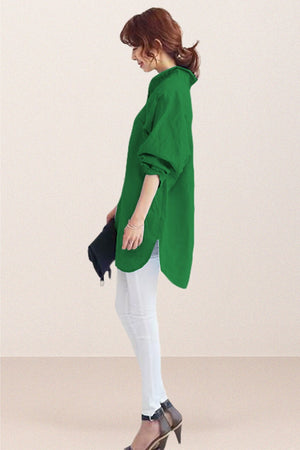 woman wearing green oversized linen shirt with rolled sleeves and white pants standing sideways holding black clutch
