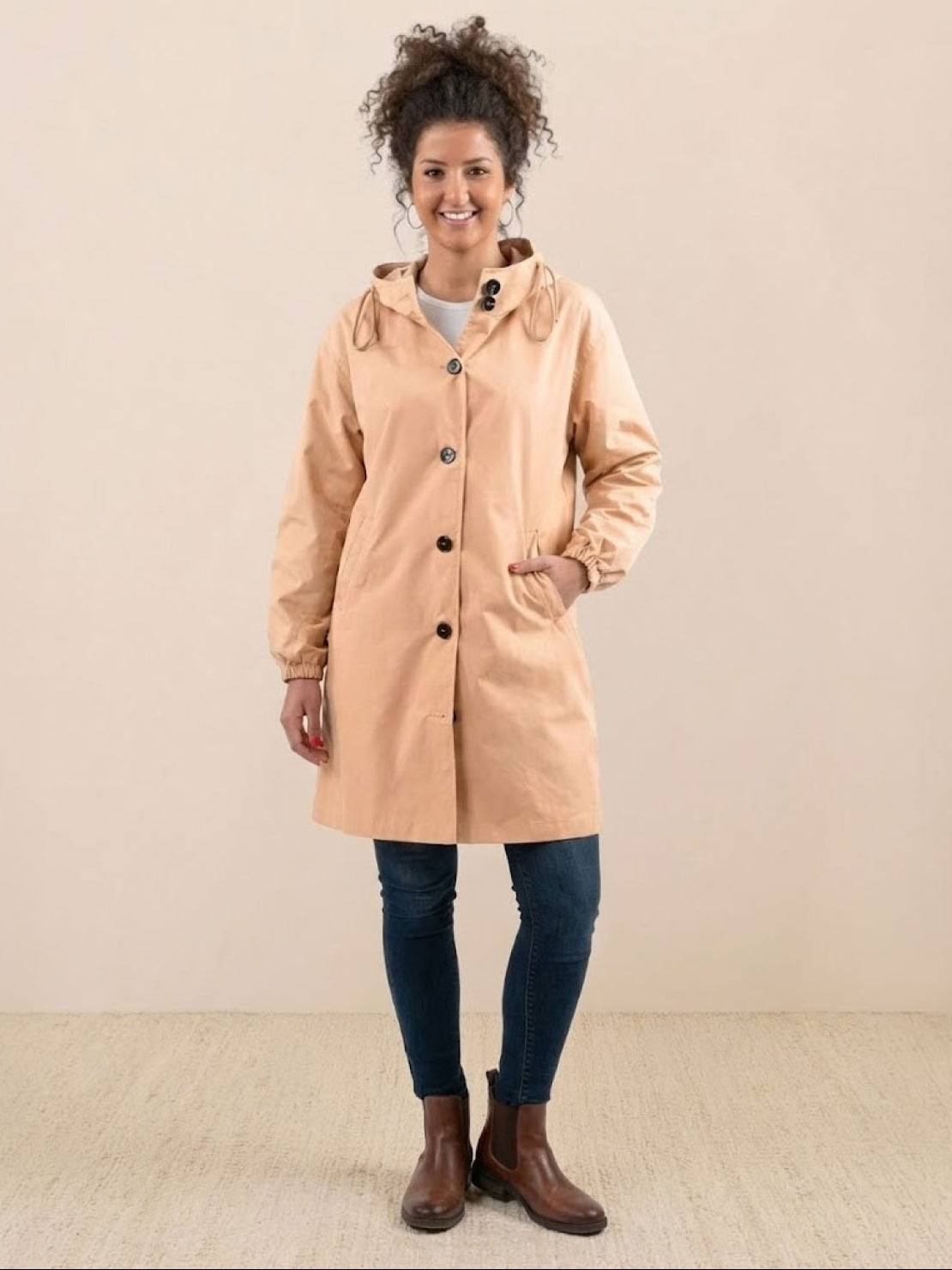 Woman wearing beige Rain Couture Windbreaker oversized water-resistant jacket with hood and pockets, paired with jeans and brown boots