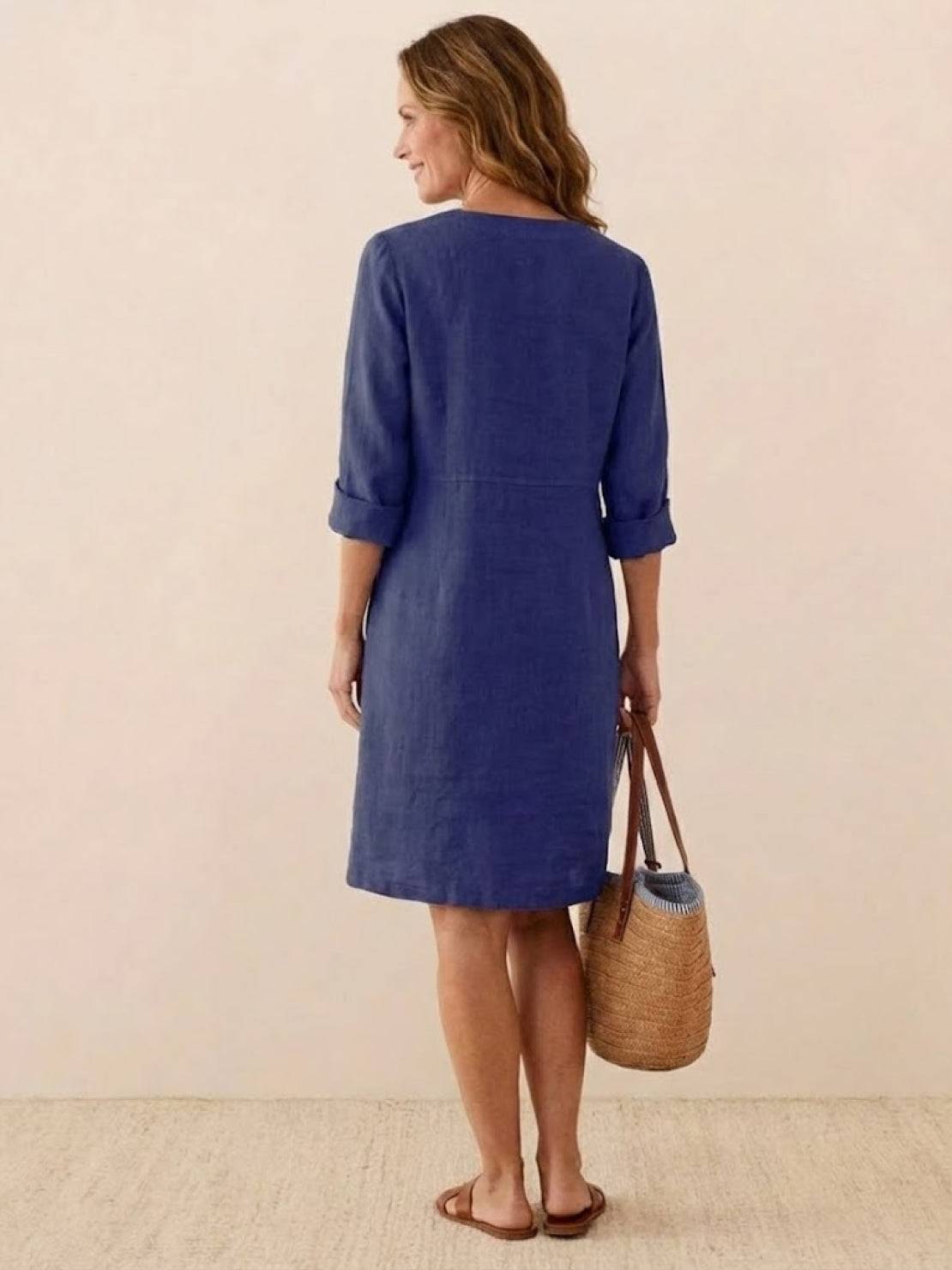 Woman wearing a blue cotton dress with rolled ¾ sleeves, holding a wicker bag, shown from the back.