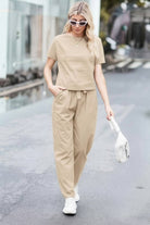 Woman wearing beige cotton linen set with short-sleeve top and straight-leg pants walking outdoors with sunglasses and white handbag