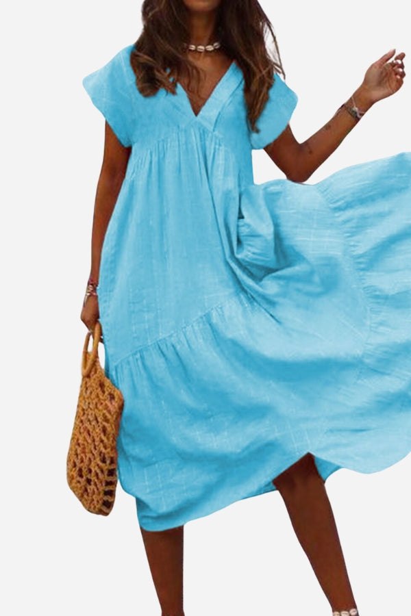Woman wearing blue V neck tiered cotton midi dress with short sleeves and frill hem, holding a woven handbag.