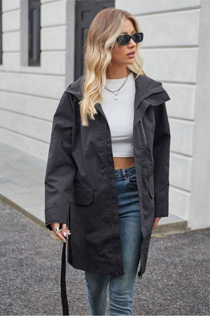 Woman wearing black water-resistant windbreaker trench jacket with belt and sunglasses outdoors