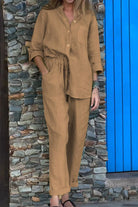 Woman wearing brown casual chic two-piece set with long sleeves and relaxed pants standing by stone wall and blue door