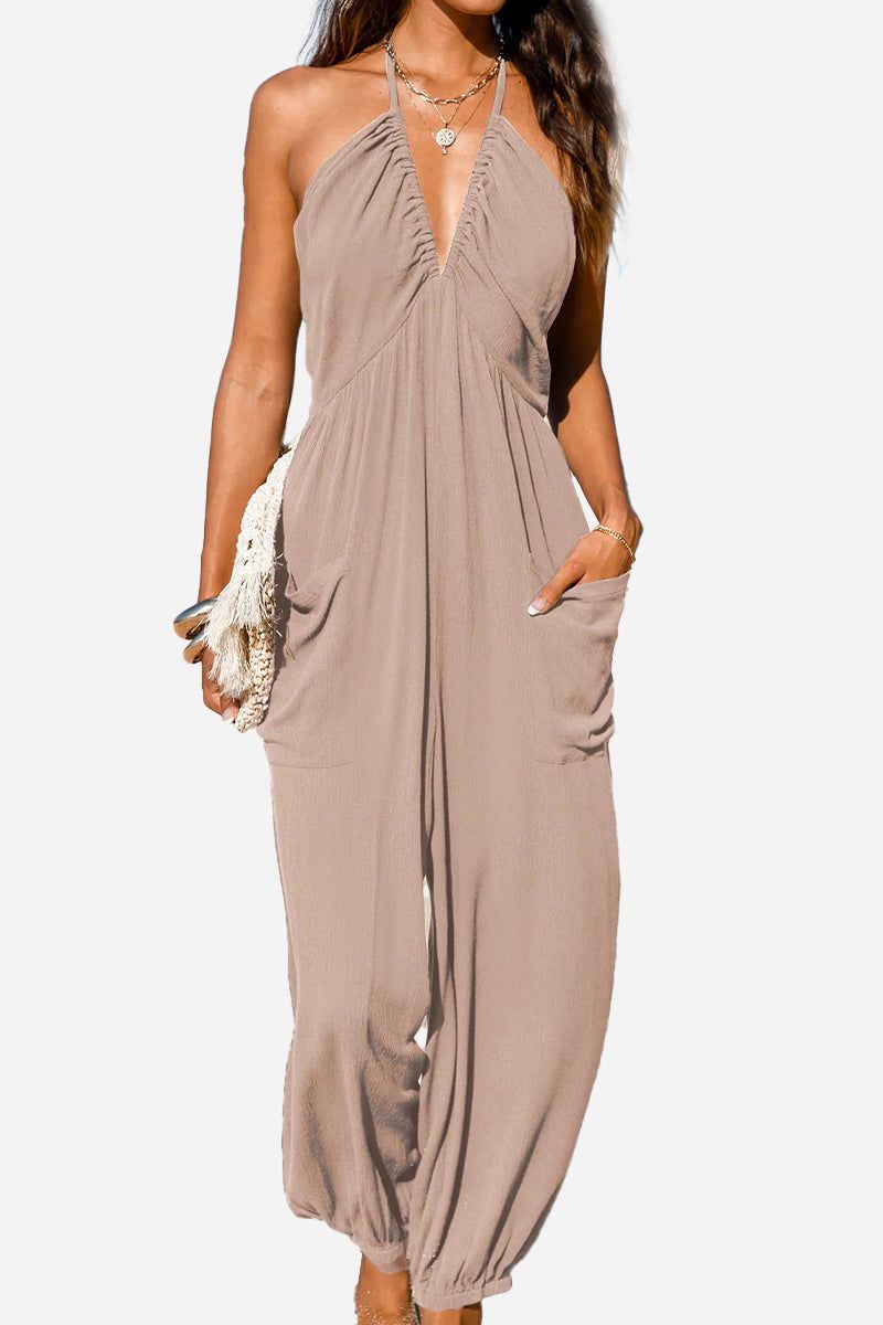 Woman wearing beige Sunshine On Sand halter-neck jumpsuit with wide legs and elastic ankle cuffs, styled with layered necklaces and holding a woven clutch bag.