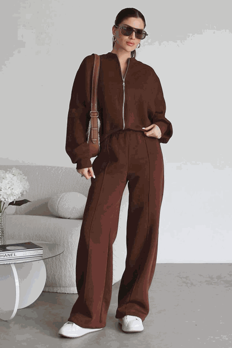 Woman wearing The Voyager Zip Set brown zip-front sweatshirt and wide-leg joggers with pockets, styled with sunglasses and white sneakers in a modern indoor setting
