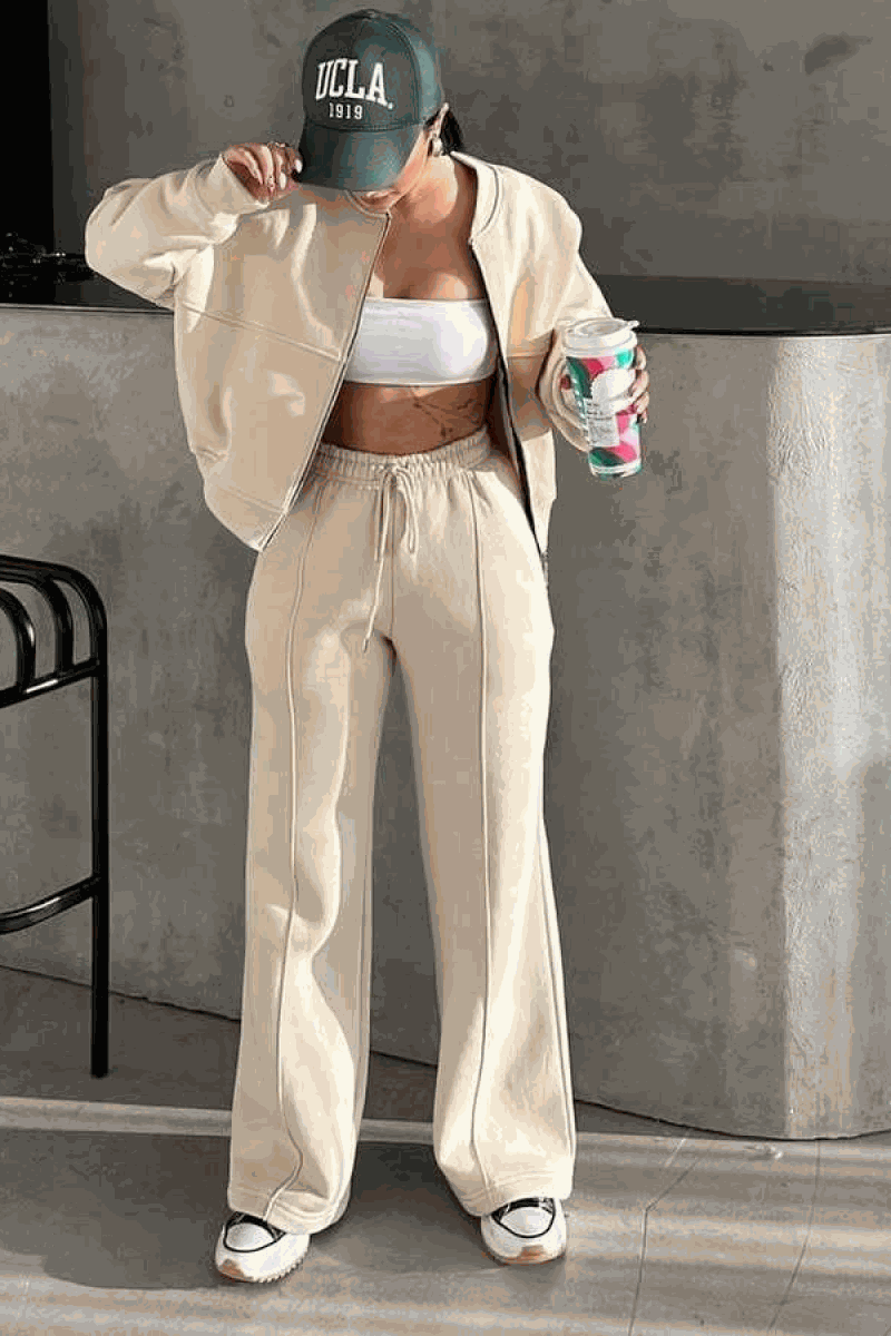 Woman wearing The Voyager Zip Set beige zip-front sweatshirt and wide-leg joggers with pockets, styled casual with sneakers and cap.