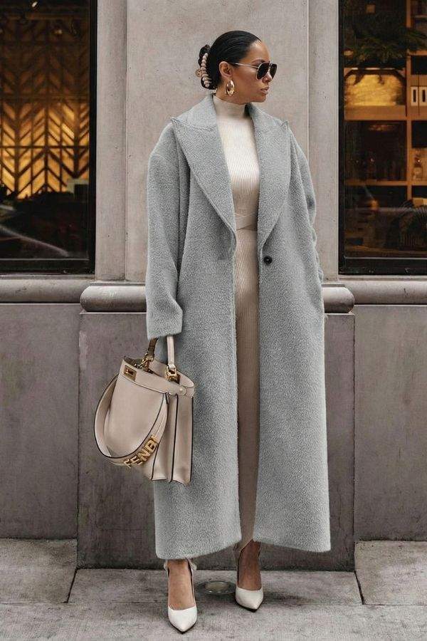Woman wearing long grey oversized teddy coat with drop shoulders and large lapels, styled with beige outfit and heels, carrying beige handbag, standing outdoors.