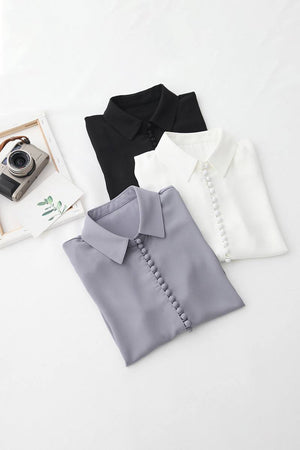Bayam Blouse folded in black, white, and gray with button details and classic collar, laid on white background with vintage camera and photo frame.