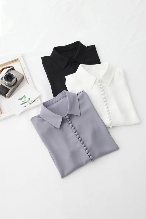 Bayam Blouse folded in black, white, and gray with button details and classic collar, laid on white background with vintage camera and photo frame.