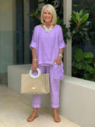 Woman wearing a lavender asymmetrical two-piece trouser set with rolled-up sleeves, paired with sandals and a beige handbag.