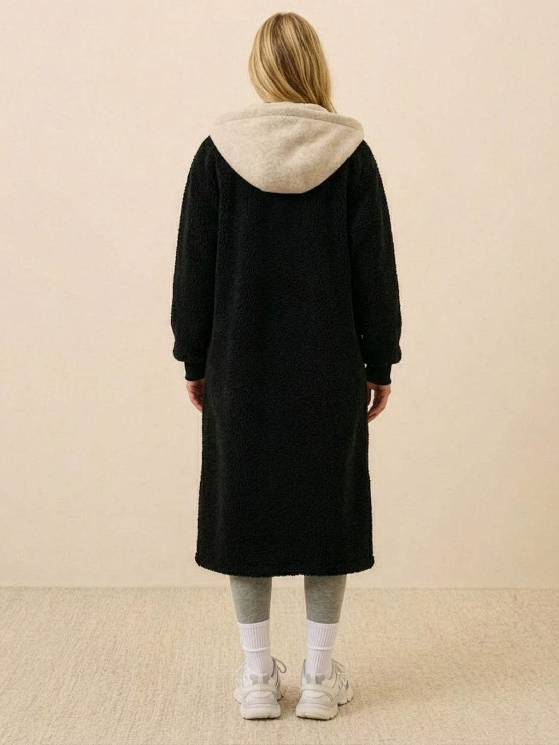 Back view of a woman wearing a black oversized sherpa long coat with a contrasting beige hood, styled casually with grey leggings and white sneakers, everyday wear oversized coat.