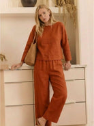 Woman wearing loose solid color rust two-piece set with long sleeves and wide-leg pants, styled casually indoors with a woven bag.