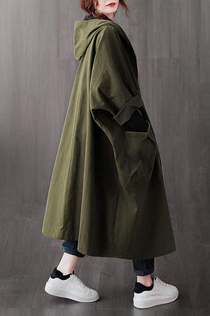Woman wearing oversized olive green water-resistant windbreaker jacket with hood and large front pockets, styled with black pants and white sneakers.