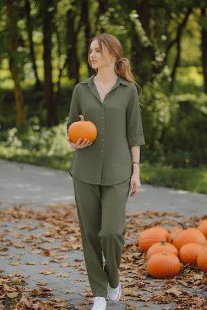Woman wearing oversized 100% cotton olive green shirt and wide-leg pants holding a pumpkin outdoors with more pumpkins on the ground.