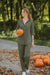 Woman wearing oversized 100% cotton olive green shirt and wide-leg pants holding a pumpkin outdoors with more pumpkins on the ground.