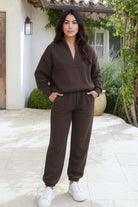 Woman wearing brown cotton blend tracksuit with zip neckline and elastic cuffs, standing outdoors in front of a house.